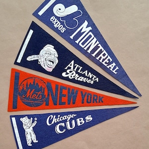 May include: Four felt pennants in blue and orange, each with team names and logos. The pennants feature the Montreal Expos, Atlanta Braves, New York Mets, and Chicago Cubs. The pennants are triangular and have white accents.