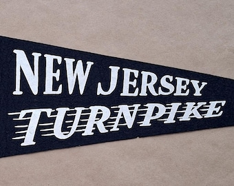 May include: A black pennant with the words "NEW JERSEY TURNPIKE" in white. The pennant features a detailed illustration of a highway interchange. The pennant is mounted on a yellow wooden frame.