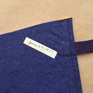 May include: A close-up of a blue fabric corner with a yellow edge and a purple strap. A white label with the handwritten text "June 25, 1957" is attached to the fabric.