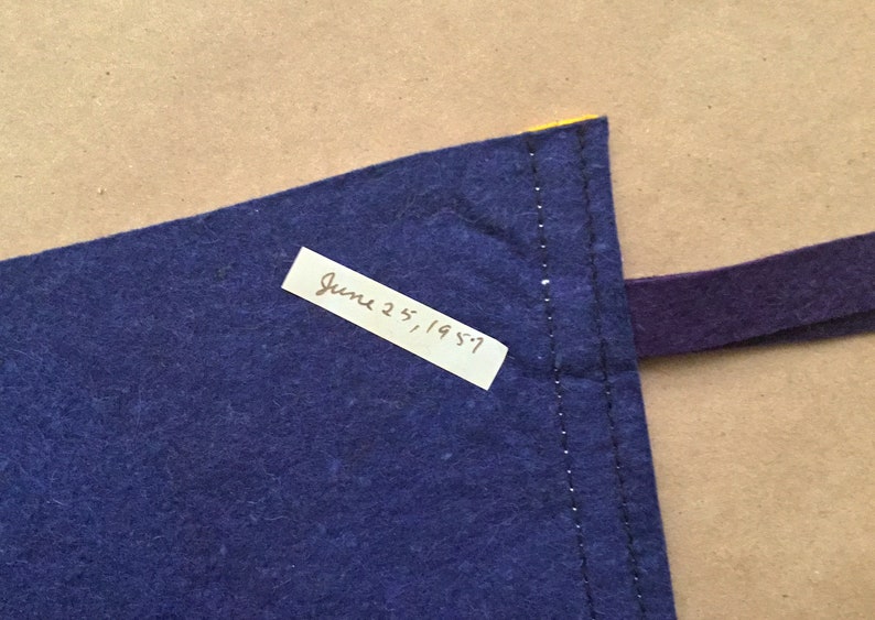 May include: A close-up of a blue fabric corner with a yellow edge and a purple strap. A white label with the handwritten text "June 25, 1957" is attached to the fabric.