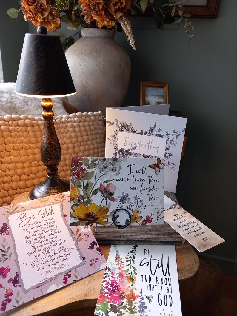 May include: A set of four sympathy cards with floral designs and inspirational quotes. The cards feature watercolour flowers in shades of pink, purple, yellow, and green. The quotes include "Be Still", "I will never leave thee, nor forsake thee", "Be Still and Know that I am God", and "Sympathy".