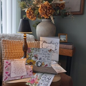 May include: A wooden table with a selection of floral greeting cards. The cards feature various floral designs and inspirational quotes. The table is decorated with a vase of dried flowers and a lamp.