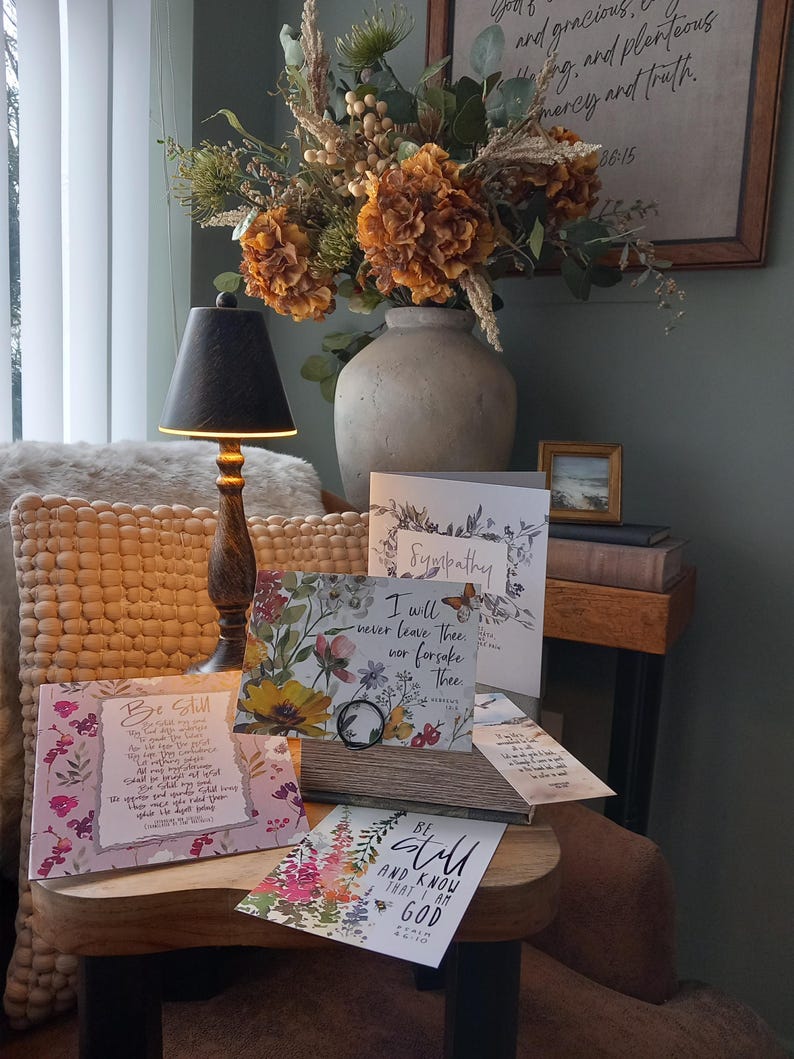 May include: A wooden table with a selection of floral greeting cards. The cards feature various floral designs and inspirational quotes. The table is decorated with a vase of dried flowers and a lamp.