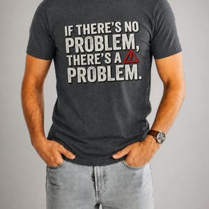 Op de afbeelding: Een antracietgrijs T-shirt met korte mouwen en de witte tekst "IF THERE'S NO PROBLEM, THERE'S A PROBLEM." met een rood-wit waarschuwingsdriehoek-icoon met een uitroepteken naast het woord "A".