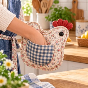 May include: A chicken-shaped oven mitt featuring a cream-colored fabric with a small pink and blue floral print. It includes a blue and white gingham pocket, a red fabric comb, a yellow beak, and a black button eye for detail.