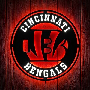Op de afbeelding: Een rond wandbord van zwart metaal met het logo van de Cincinnati Bengals en de teamnaam in witte letters. Het ontwerp wordt van achteren verlicht met een levendige rode gloed, waardoor de uitgesneden details van de tijgerstrepen opvallen.