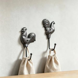Könnte beinhalten: Zwei dunkelgraue Wandhaken aus Metall in Hahnform, jeweils mit strukturierter Oberfläche und einem gebogenen Haken am unteren Ende. An jedem der beiden hahnförmigen Wandhaken hängt ein kleiner, beigefarbener Stoffbeutel mit Kordelzug.