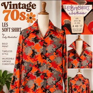 May include: A long-sleeve button-down shirt featuring a bold, abstract geometric pattern in vibrant orange, black, and grey. The shirt has a pointed collar, a front button placket, and a label reading Les Soft Shirt by Lady Manhattan.