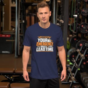 以下が含まれることがあります： 白とオレンジのグラフィックプリントが施されたネイビーブルーの半袖Tシャツ。胸の中央には「LOGISTICS YOUR EMERGENCY HAS A LEAD TIME」というテキストと、小さな時計のアイコン、トラックのイラストが描かれています。