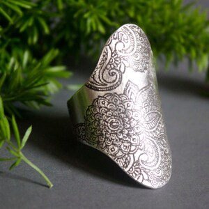 Könnte beinhalten: Ein silberner Ring mit einem filigranen Blumendesign. Der Ring ist tropfenförmig und hat ein breites Band.