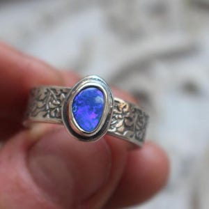 Puede incluir: Un anillo de plata con una piedra de ópalo azul ovalada engastada en un engaste de bisel. La banda está decorada con un diseño floral.