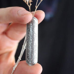 May include: A silver pendant necklace featuring a floral design. The rectangular pendant is intricately carved with a repeating pattern of flowers and leaves. A delicate silver chain is attached. A small dried flower stem is inserted into the pendant.