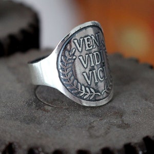 May include: A silver ring with a laurel wreath design and the inscription "VENI VIDI VICI" on the face.