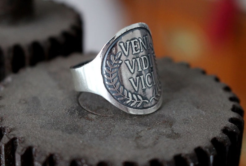 May include: A silver ring with a laurel wreath design and the inscription "VENI VIDI VICI" on the face.