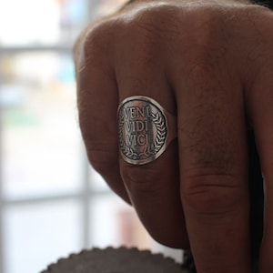 May include: A silver ring with a round face featuring the Latin phrase "veni vidi vici" surrounded by a laurel wreath.