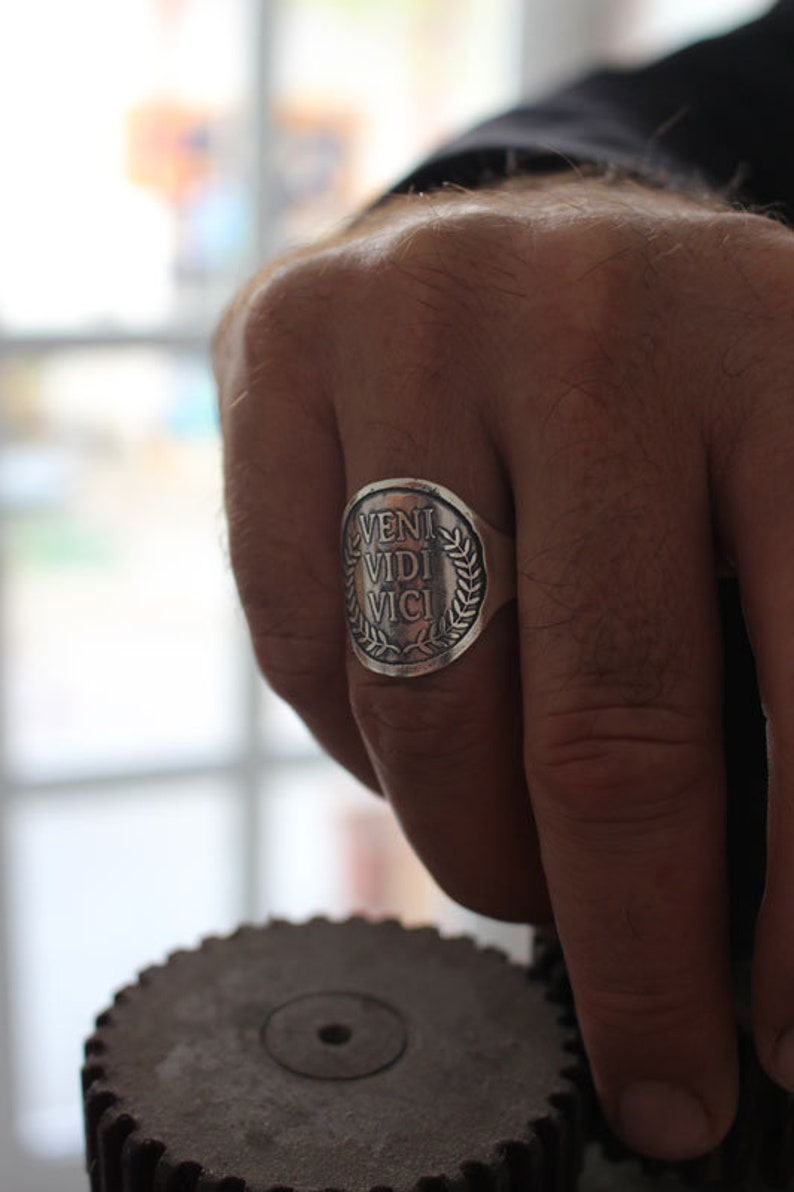 May include: A silver ring with a round face featuring the Latin phrase "veni vidi vici" surrounded by a laurel wreath.