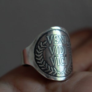 May include: A silver ring with a raised circular design featuring the Latin phrase "veni vidi vici" surrounded by a laurel wreath.