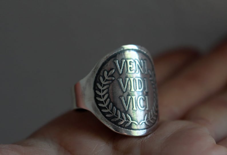 May include: A silver ring with a raised circular design featuring the Latin phrase "veni vidi vici" surrounded by a laurel wreath.
