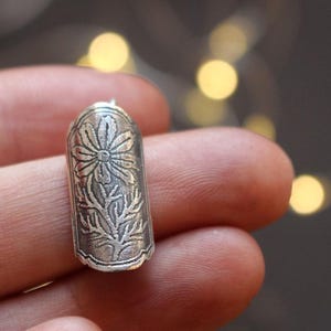 May include: A silver-colored thimble with an embossed floral design. The thimble is cylindrical with a rounded top and features a detailed pattern of a flower and leaves. The thimble is held between fingers.