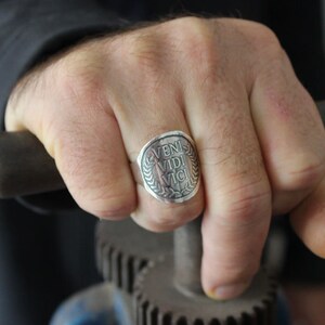 May include: A silver ring with a round face featuring the Latin phrase "veni vidi vici" surrounded by a laurel wreath.