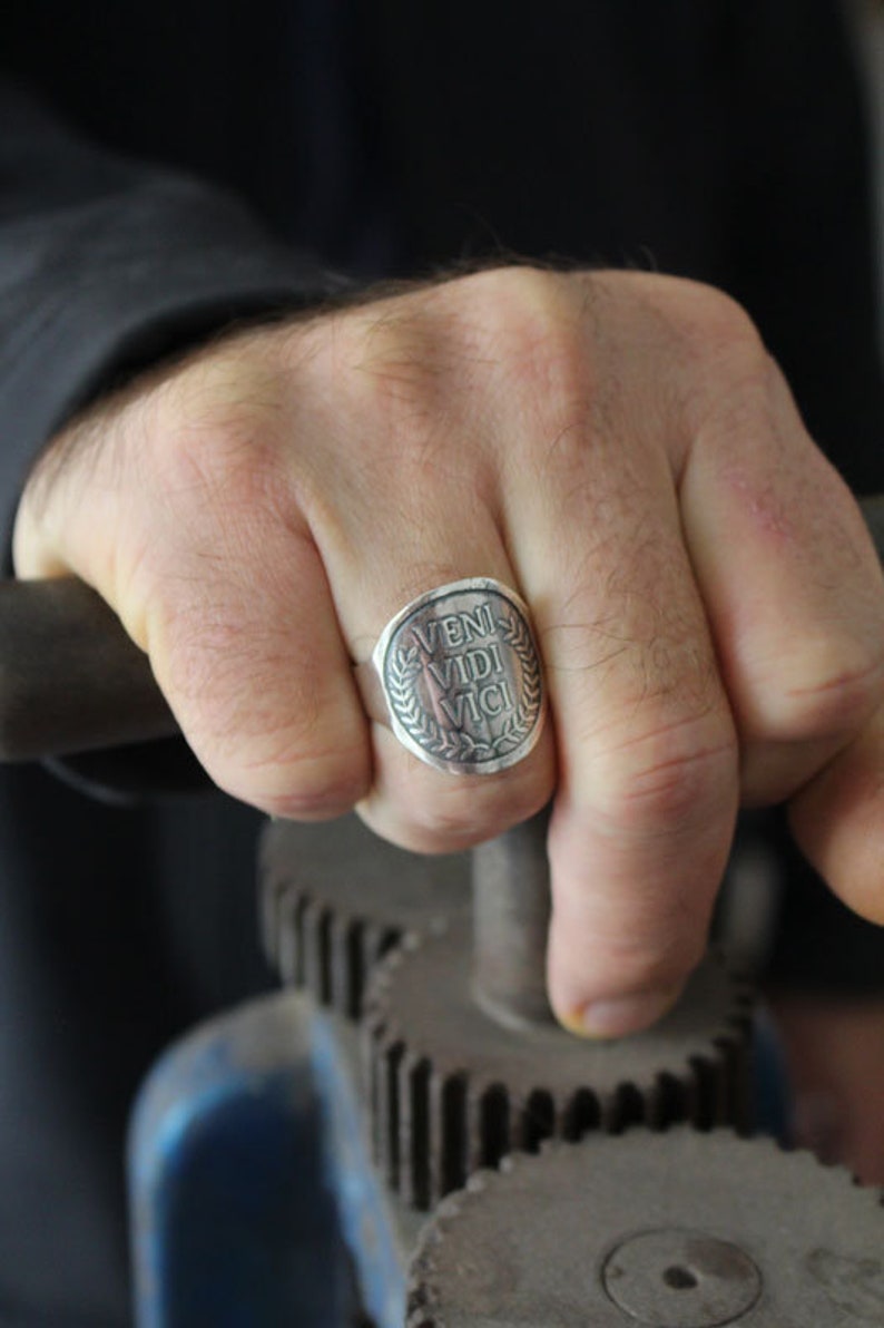 May include: A silver ring with a round face featuring the Latin phrase "veni vidi vici" surrounded by a laurel wreath.