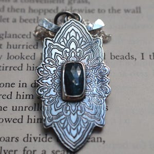 Puede incluir: Colgante plateado con un diseño floral ornamentado y una piedra preciosa rectangular de color azul oscuro. El colgante está sobre una cadena y sobre un fondo de texto impreso. Se destacan los intrincados detalles del colgante.