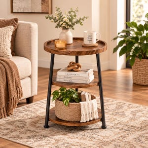 May include: A three-tier round side table with a dark wood grain finish and black metal legs. The top shelf holds a vase with greenery, a candle, and a white mug with the text Good Things Ahead. The middle shelf holds books and a knot decor.
