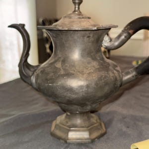 May include: A vintage pewter teapot featuring a bulbous body, a curved spout, and a dark wooden handle. The teapot has a removable lid with a small finial knob and rests on an octagonal pedestal base. The metal surface shows a dull, aged patina.