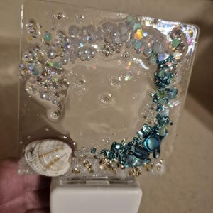 May include: A square night light featuring a clear resin panel embedded with iridescent glitter, small white beads, and a curved arrangement of blue and teal crushed stones. A single white seashell is positioned at the bottom left corner.