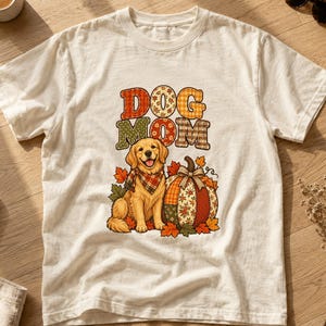 Könnte beinhalten: Ein cremefarbenes Kurzarm-T-Shirt mit einem Grafikdruck eines Golden Retrievers, der ein kariertes Halstuch trägt, neben einem Patchwork-Kürbis. Der Text DOG MOM ist in bunten, gemusterten Blockbuchstaben mit Herbstblättern darüber gedruckt.