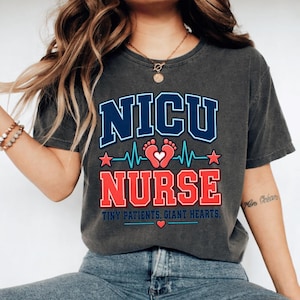 Könnte beinhalten: Ein anthrazitfarbenes Kurzarm-T-Shirt mit dem Text NICU NURSE in blauer und roter Schrift. Das Design enthält eine hellblaue Herzschlaglinie, ein Paar rote Babyfußabdrücke mit einem weißen Herz und den Text TINY PATIENTS. GIANT HEARTS.