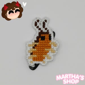 May include: A cross-stitch patch shaped like a cockroach with a brown body, black eyes, and light tan legs, outlined in white thread. The insect figure is attached to a black metal safety pin. In the top left corner is a cartoon character icon.