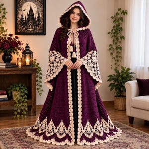 May include: A floor-length, deep purple crochet hooded cloak featuring intricate cream-colored lace trim along the hood, wide sleeves, and hem. The garment includes a front tie closure with two decorative tassels, showcasing a detailed stitch pattern.