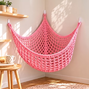 May include: A bright pink crocheted corner hammock featuring a diamond-shaped net pattern. The hammock is suspended from three white adhesive hooks attached to the corner of the walls, creating a triangular storage space for various items.