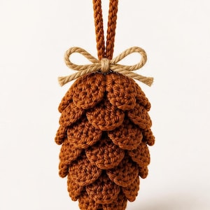 May include: A crocheted pinecone ornament in a rich brown yarn, featuring layered, textured scales. The ornament is topped with a small, light beige jute twine bow and a matching braided loop for hanging, creating a detailed, handcrafted look.