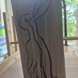May include: A book art sculpture created by folding the pages of a book to form the silhouette of a standing hare with long ears. The white paper edges are precisely manipulated to create the outline of the animal, showing a side profile view.