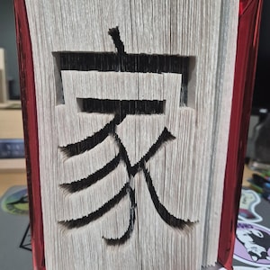 May include: A book art sculpture featuring the Chinese character for home, 家, created by folding individual pages. The book has a metallic red cover visible on the sides, and the folded edges of the white paper pages form the black-inked character.