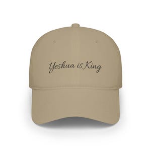 May include: A tan baseball cap featuring the text Yeshua is King embroidered in black script font across the front panels. The cap has a structured crown, a curved brim, and a button at the top centre, providing a clean and simple design.