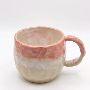Pode incluir: Uma caneca de cerâmica feita à mão com um corpo arredondado e irregular e uma pega grossa. O exterior apresenta um esmalte de dois tons com uma secção superior rosa suave que transita para uma base creme, com o interior branco brilhante.