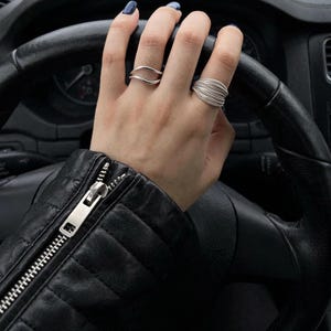 Könnte beinhalten: Zwei Silberringe an den Fingern. Ein Ring hat ein schlichtes, glattes Design in Form einer offenen Welle. Der zweite Ring besteht aus mehreren dünnen, parallelen Silberdrähten, die zu einem breiten, strukturierten Band mit Schichtoptik gewickelt sind.