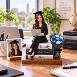 May include: A figurine of a graduate in a black suit and cap sitting on a stack of books labelled Emily, Software Engineer, and 2026, holding a laptop. Beside it is a bouquet of blue and white orchids, a framed photo, and a pen holder with text.