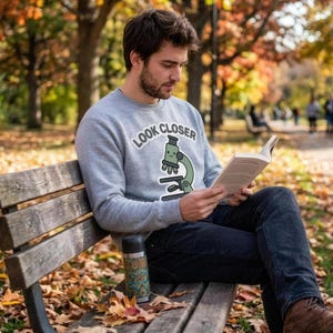 May include: A light gray crewneck sweatshirt featuring a graphic of a green microscope with a smiling face and the text "LOOK CLOSER" printed in an arc above it, paired with dark blue denim jeans and brown leather boots.