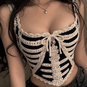 May include: A black and cream crochet corset top featuring a rib cage pattern in cream yarn against a black background. The garment has a sweetheart neckline, a central tie closure with a small silver heart charm, and a scalloped edge finish.