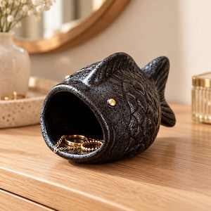 May include: A black ceramic fish-shaped trinket dish with a textured, speckled finish and a wide, open mouth. The interior of the fish holds two gold-toned metal rings and a delicate gold chain, while small gold studs serve as the fish's eyes.