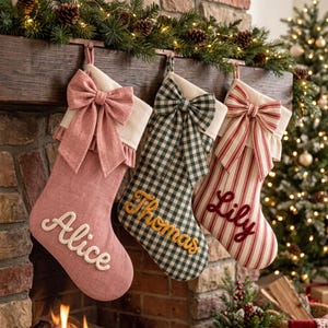 May include: Three Christmas stockings hanging in a row. The first is solid dusty pink with the name Alice in white script. The second is green and white gingham with the name Thomas in yellow. The third is red and white striped with the name Lily in red.