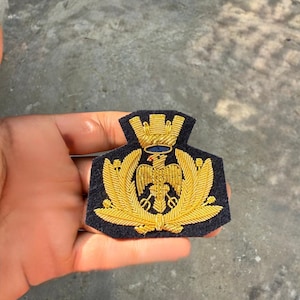 May include: A dark navy blue embroidered patch featuring a gold metallic thread design of an eagle centred within a laurel wreath, topped with a mural crown. The intricate needlework highlights the bird's wings and the crown's architectural details.