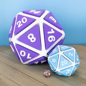 May include: A large purple twenty-sided die with white numbers and a white frame, a medium light blue twenty-sided die with white numbers and a white frame, and a small metallic pink twenty-sided die with black numbers sitting together.