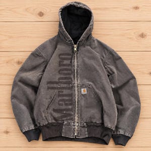 Peut inclure: Une veste à capuche gris anthracite avec une fermeture éclair intégrale, présentant le mot Marlboro imprimé verticalement en police foncée sur le côté droit et un petit écusson Carhartt sur la poche poitrine gauche, avec poignets et ourlet côtelés noirs.