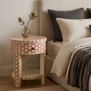 May include: A round bedside table with a cream-coloured base and a repeating geometric pattern in reddish-brown. The table features a single drawer with a small dark knob and an open lower shelf. A clear glass vase with a plant sits on top.