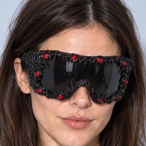Puede incluir: Gafas protectoras negras de gran tamaño con una montura texturizada y rugosa, adornada con pequeñas piedras preciosas rojas circulares incrustadas a lo largo del borde. Las lentes son oscuras y reflectantes, cubriendo completamente el área ocular.